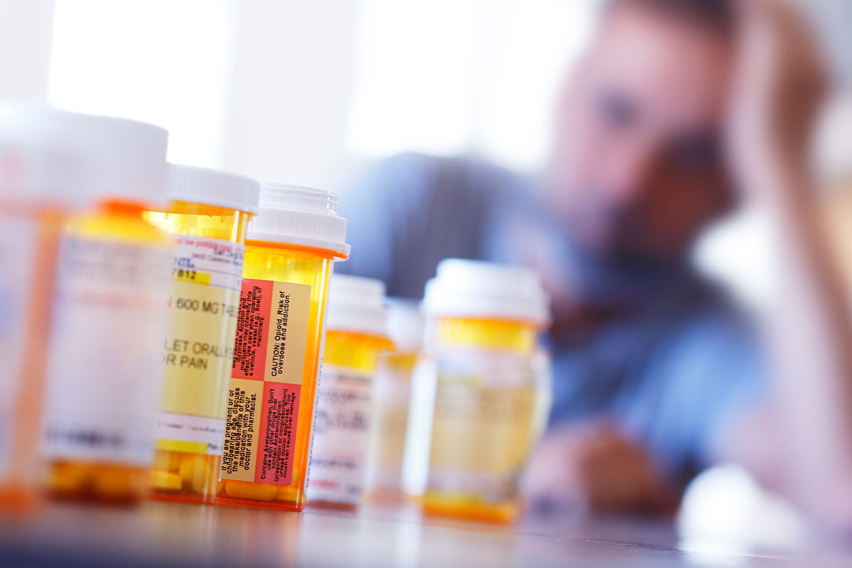 Opioid abuse concept with prescription opioid pill bottles in foreground and out-of-focus man in background. Get the latest updates on America’s opioid crisis for 2022.