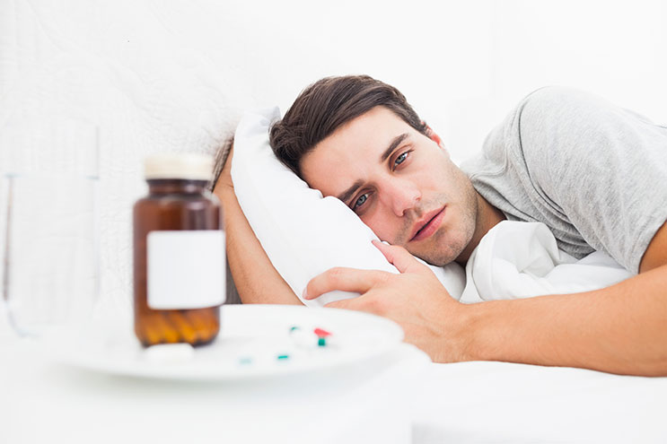 A tired-looking man lies in bed staring at pills on his nightstand.