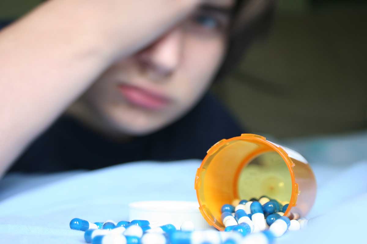 An image conveying a depressed teen boy with a bottle of prescription medications. An rx pill bottle is in focus in the foreground with a blurred sad teenage boy in the background.