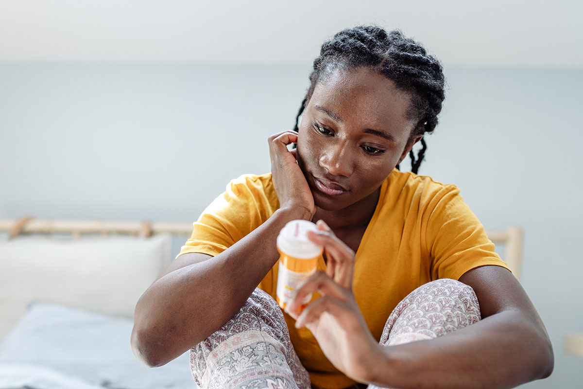 A young woman sitting on her bed in her room, looking at a prescription bottle and considering misusing an opioid prescription.