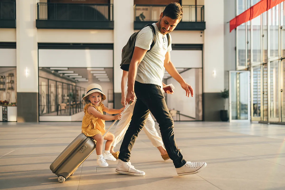 A family travels through an airport, a smiling toddler riding on the top of her father’s rolling carry-on suitcase. Learn how to protect your traveling family with the Rx Locking Bag.